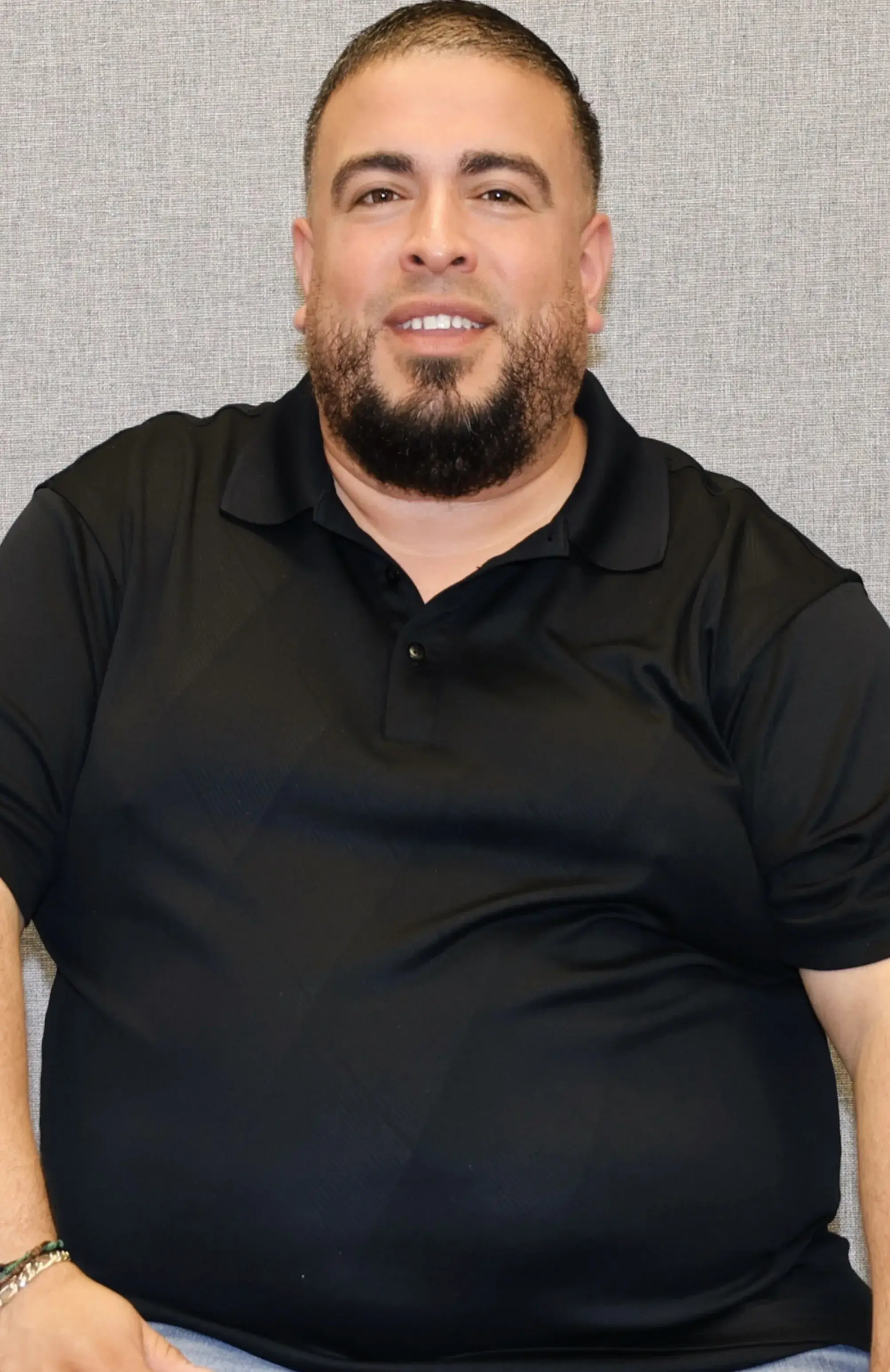 Man in a black polo shirt sitting against a gray background.