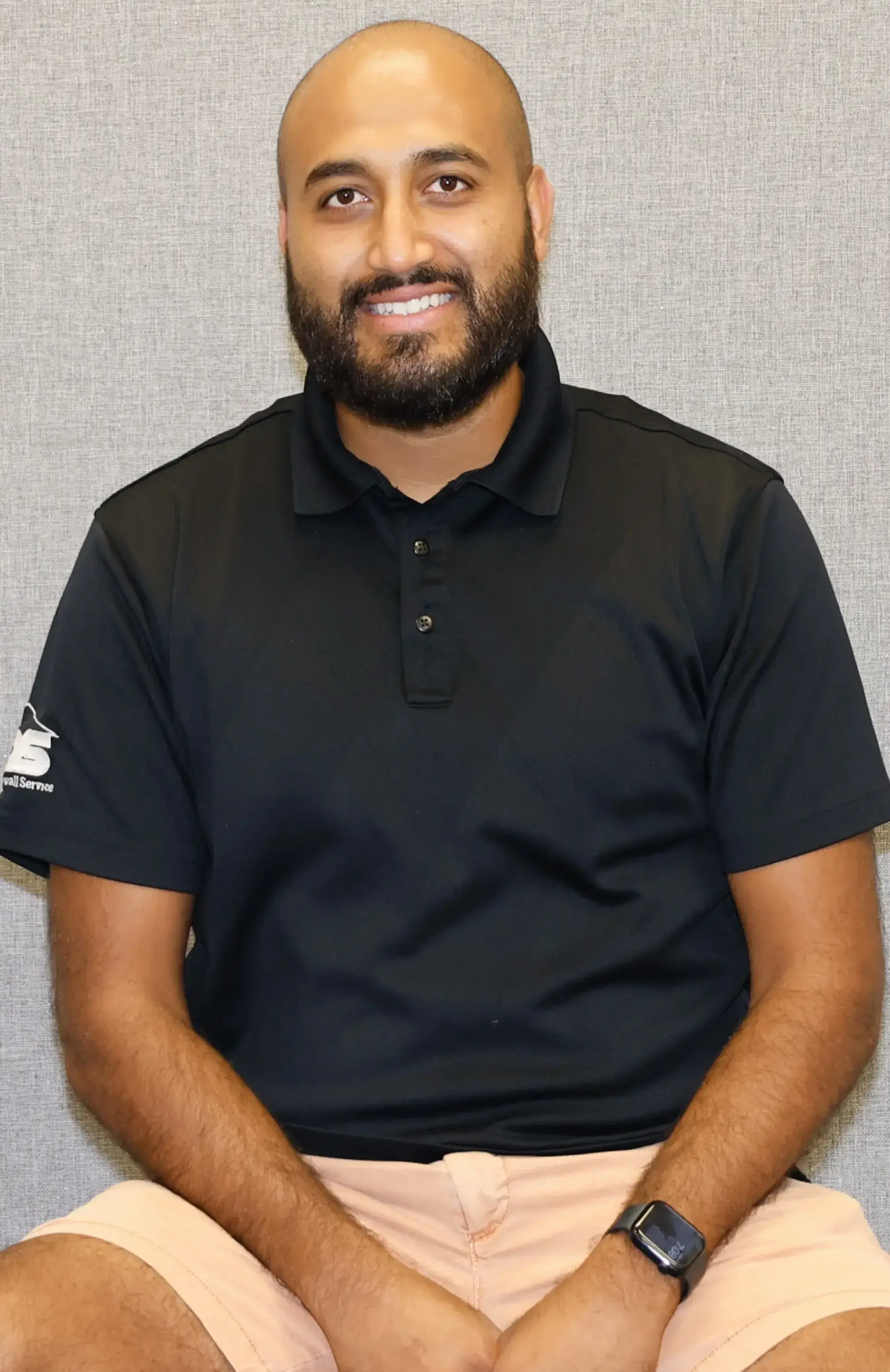 Man smiling, wearing a black polo shirt, sitting against a gray background.