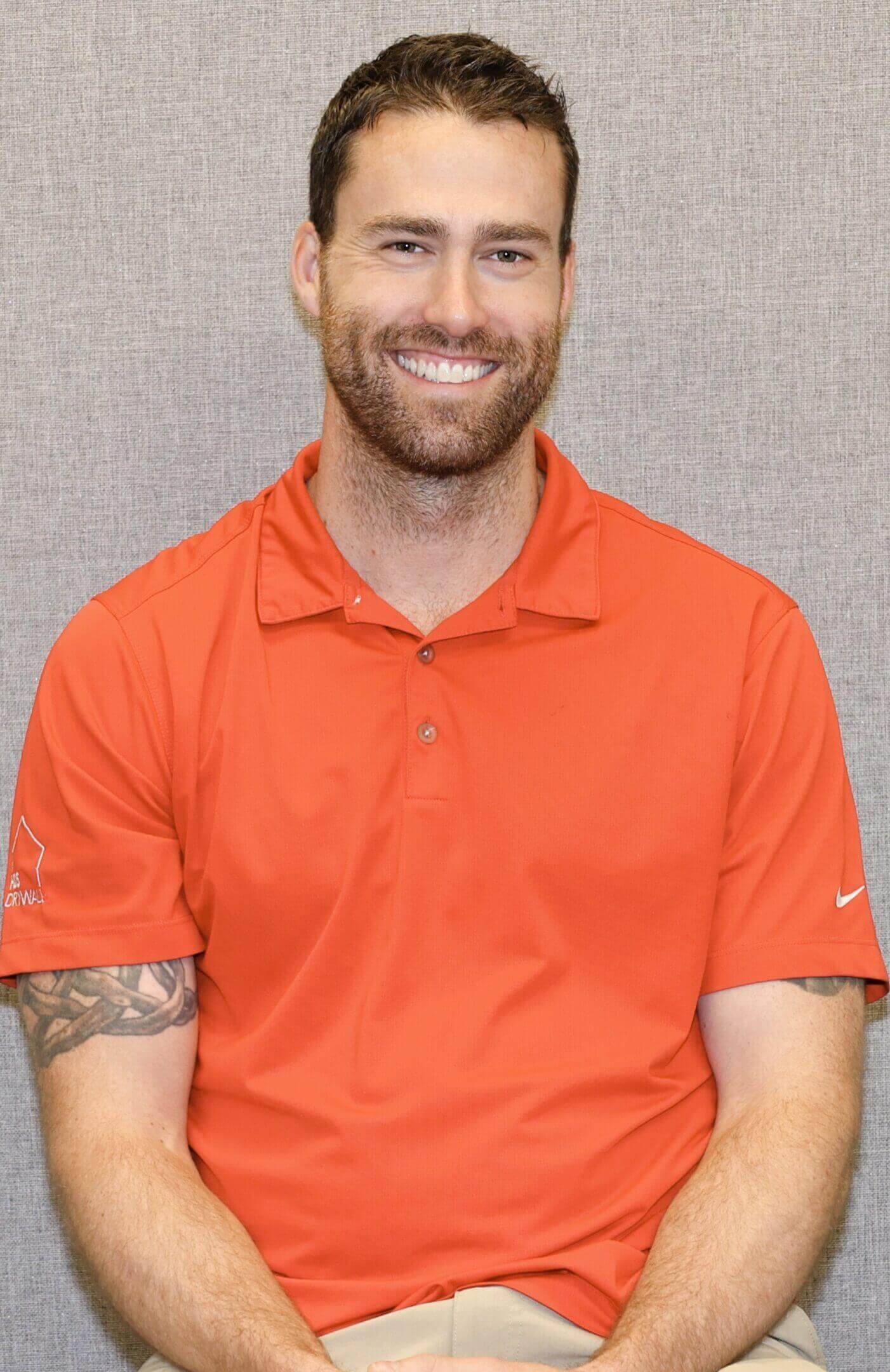 Smiling man in an orange polo shirt with tattoos on his arms.